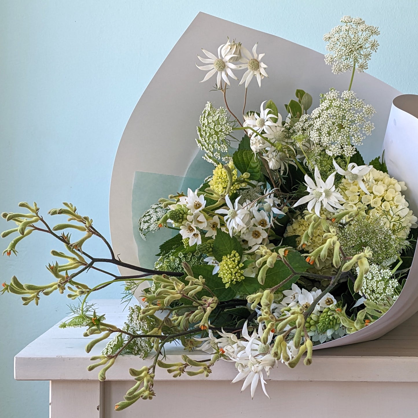 Front view of a bouquet of white, yellow and green native Australian flowers wrapped in white paper, lying on a white table with a blue background.
