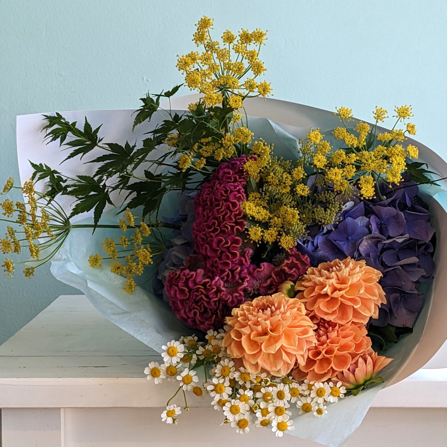 Front view of a seasonal flower bouquet in white, yellow, orange, green and blue. The bouquet is wrapped in white paper and lying on a white table with a blue background.