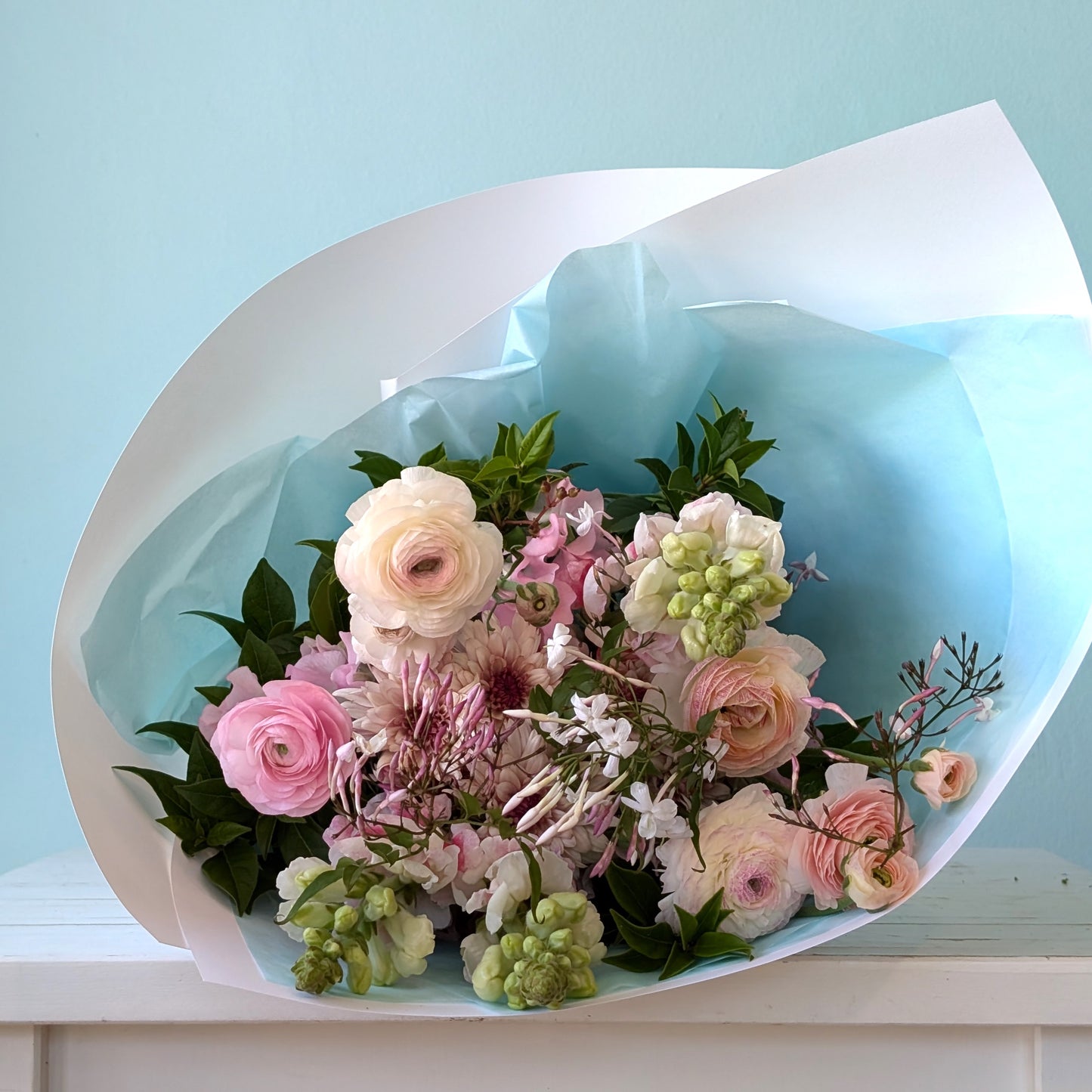 Front view of a small posy of flowers in soft pink, white, green and purple, wrapped in white paper and lying on a white table with a blue background.