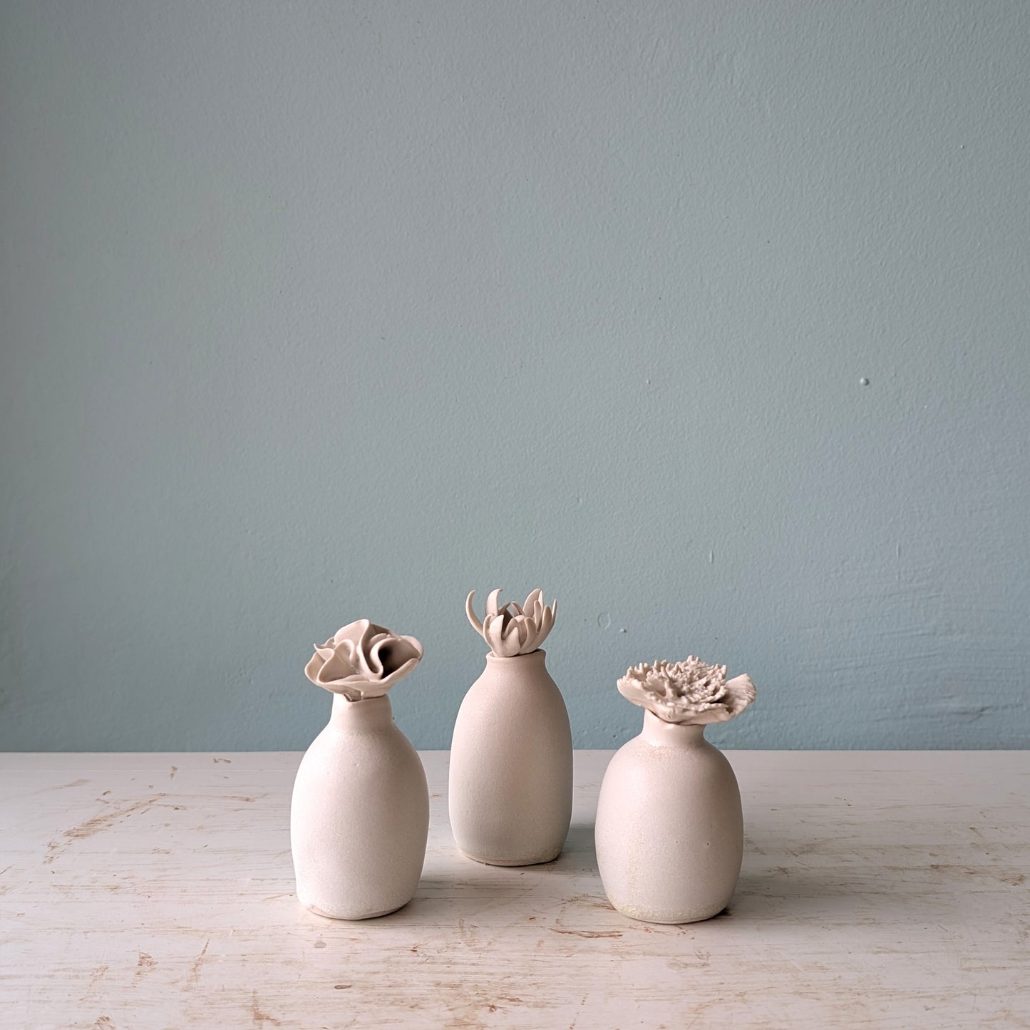 Three small matte ceramic vases in white, arranged on a light wooden surface against a pale blue wall.
