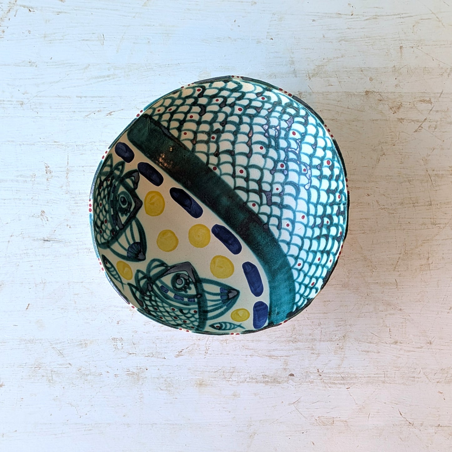A hand-painted ceramic bowl with a white base, featuring a green fish-scale pattern, dark green fish motifs, and yellow polka dots, shown from a top-down view on a distressed white wooden surface.