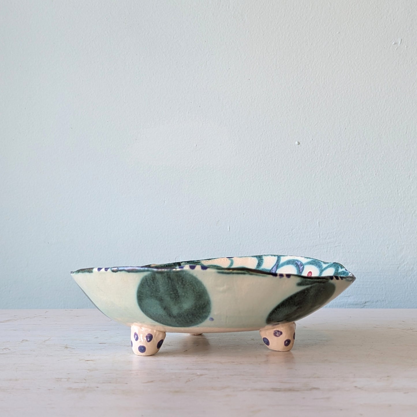 A handmade ceramic footed bowl with a cream base, dark green and blue-green painted spots, and purple polka-dot feet, set against a light blue wall.