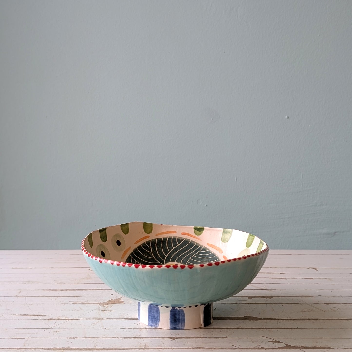 Front view of a colourful ceramic bowl with a foot ring, on a white wooden surface with a blue background.