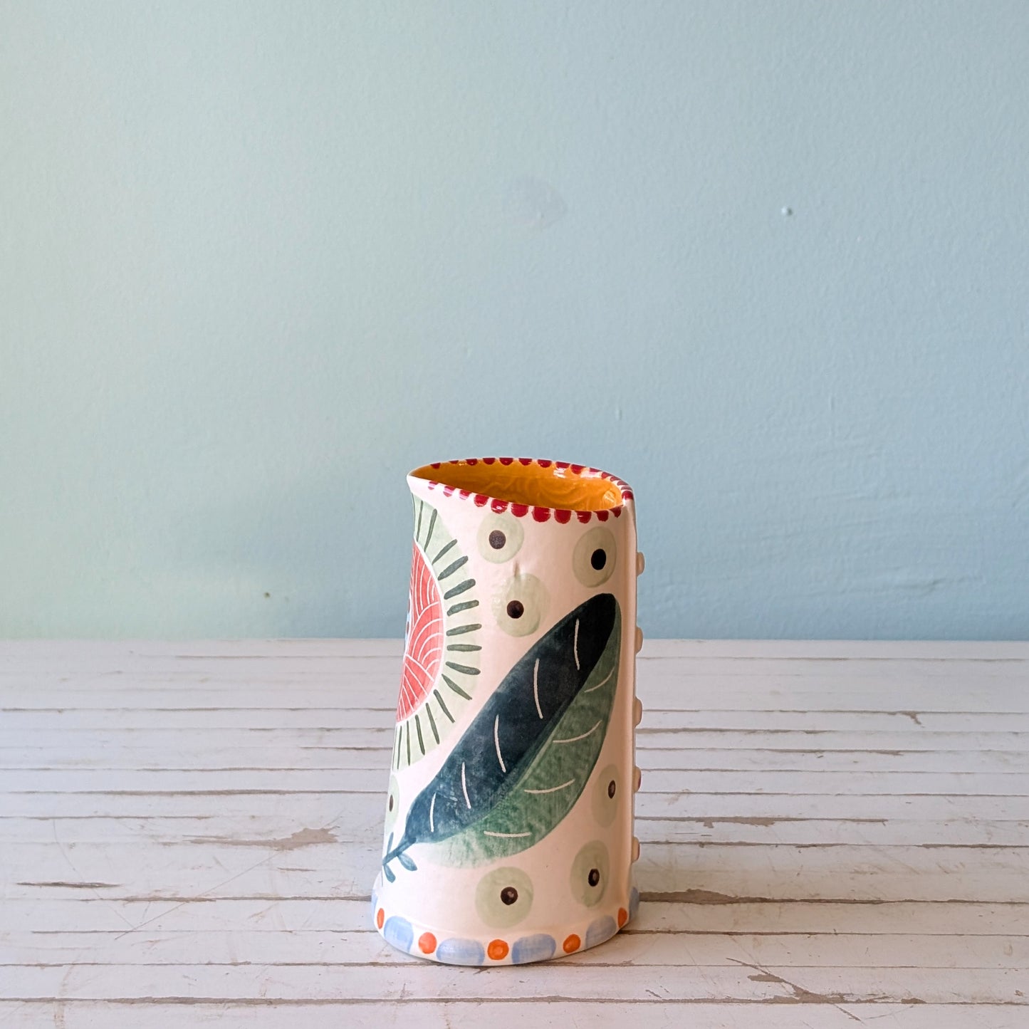 Front view of a white ceramic pourer with green, blue and orange botanical pattern and orange exterior