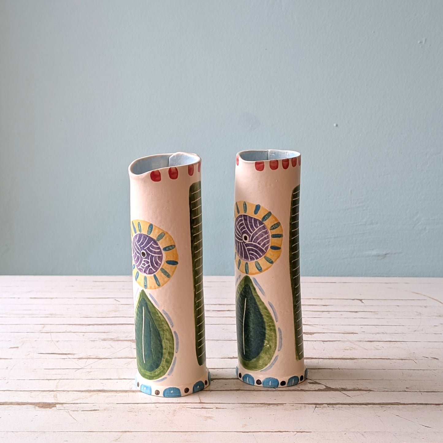 Front view of two colourful ceramic pourers on a white wooden surface against a blue background.