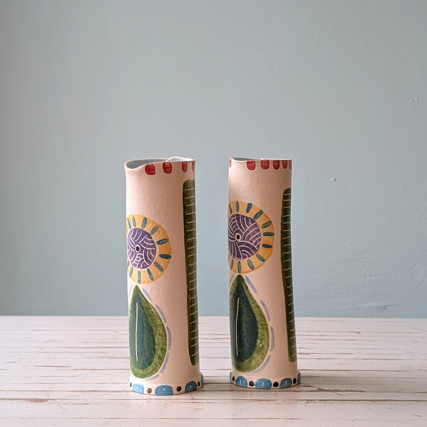 Front view of two colourful ceramic pourers on a white wooden surface against a blue background.