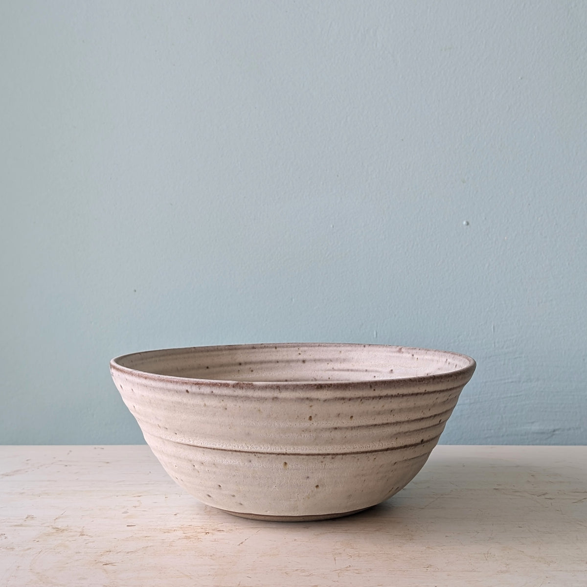 A handmade ceramic bowl with a textured, ribbed exterior and a rustic speckled finish, sitting on a light wooden surface against a muted blue wall.