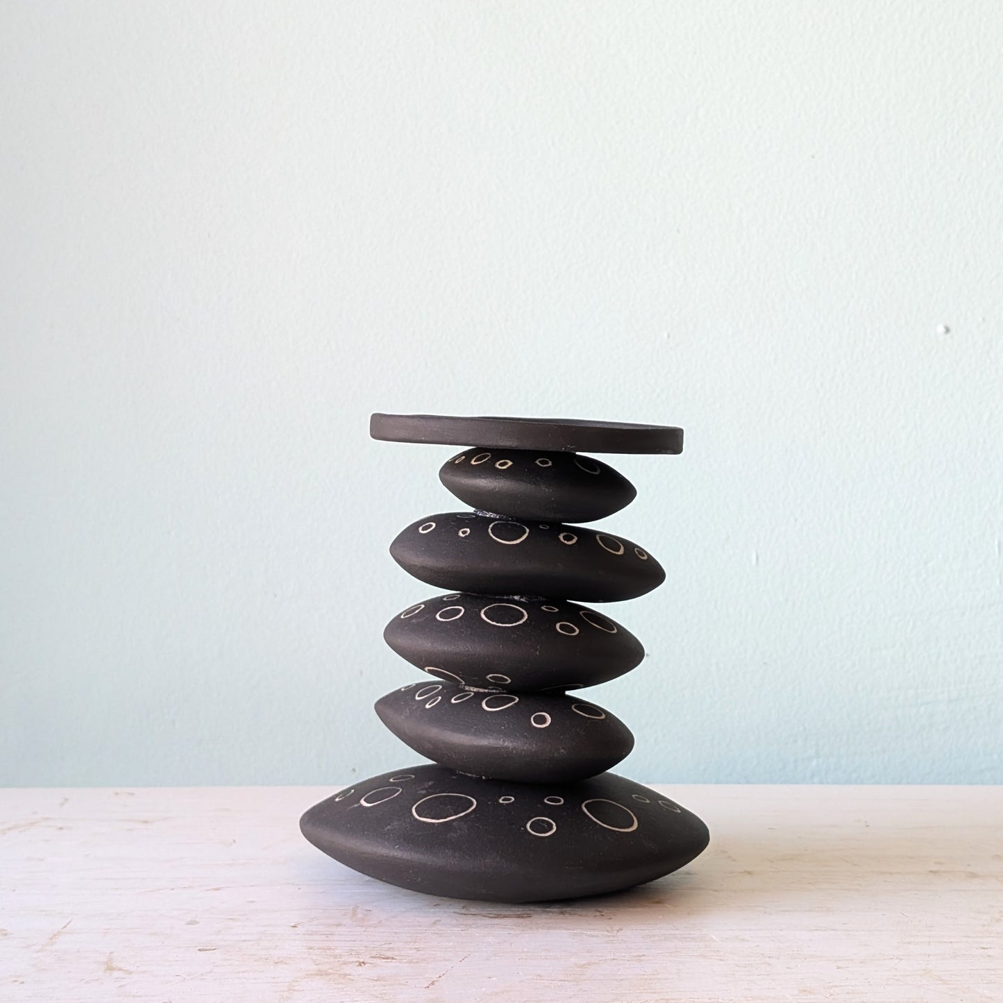 A black ceramic stack candlestick with white hand-drawn circle details, displayed on a light-colored wooden surface against a pale blue wall.