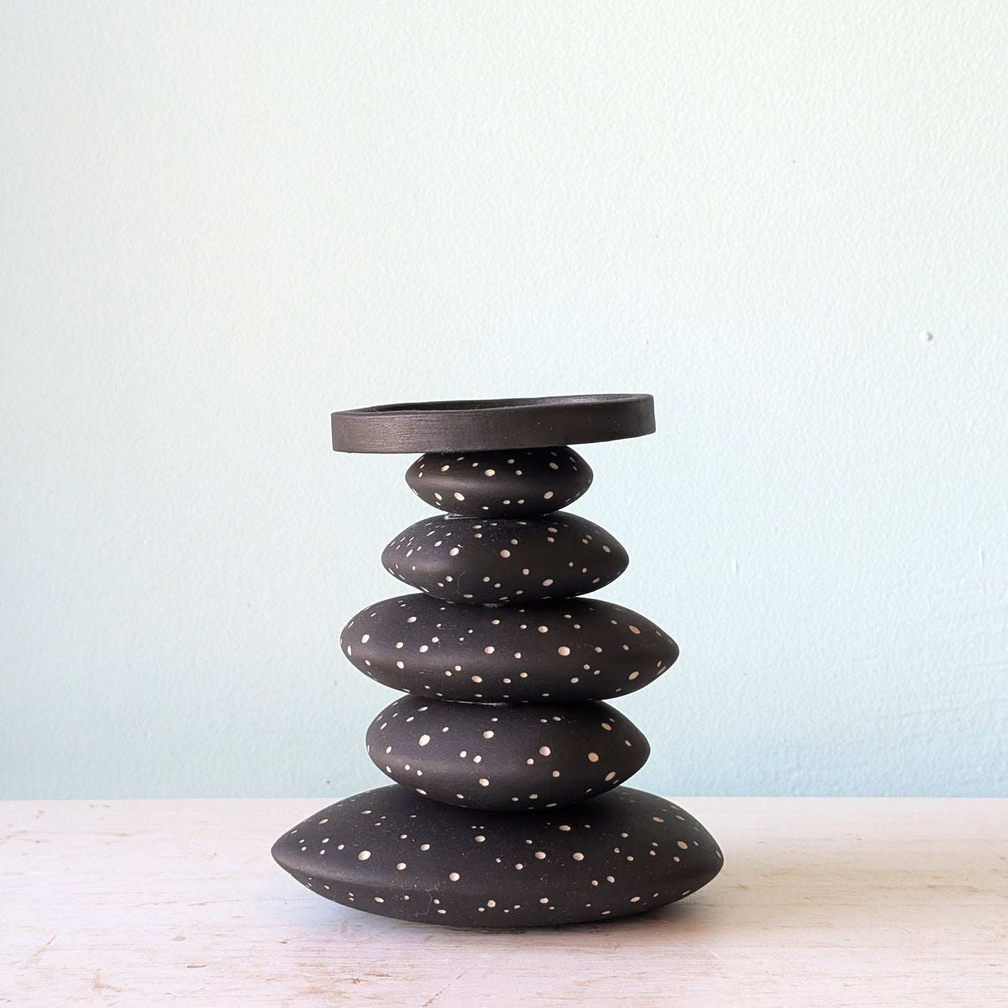 A black ceramic stack candlestick with white hand-drawn dot details, displayed on a light-colored wooden surface against a pale blue wall.