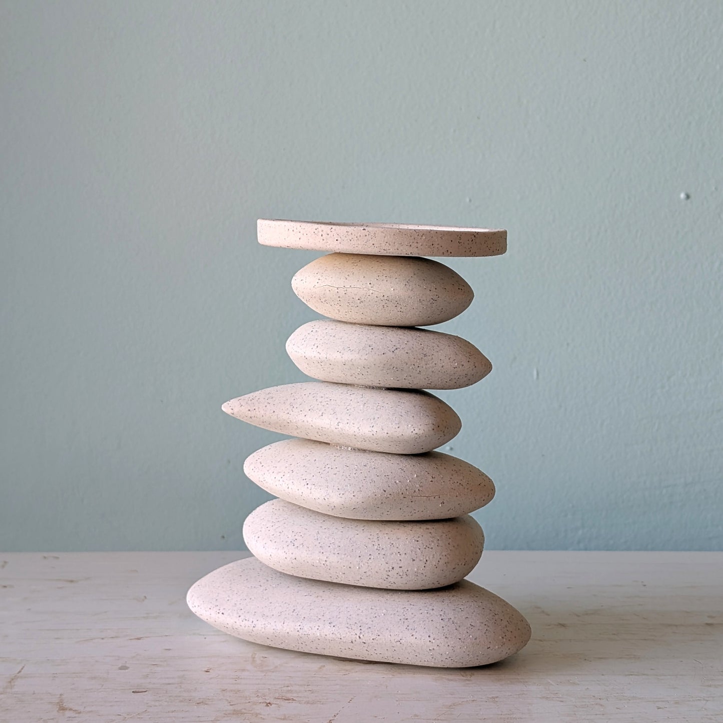 A candlestick in that shape of a stack of organic-shaped ceramic stones in speckled cream-beige, on a light wooden surface against a blue wall.