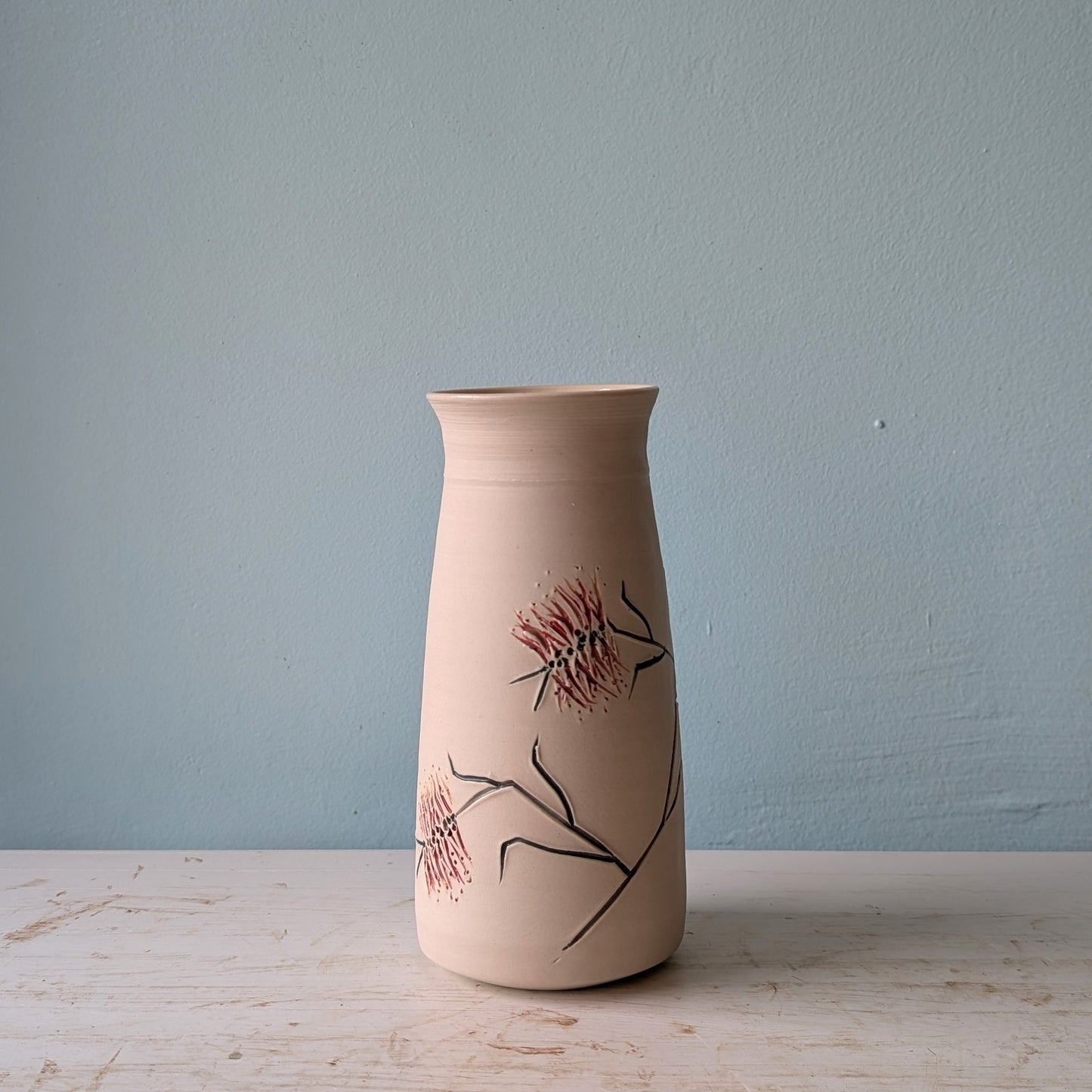 Ceramic vase with floral bottlebrush design on a white table against a blue wall.