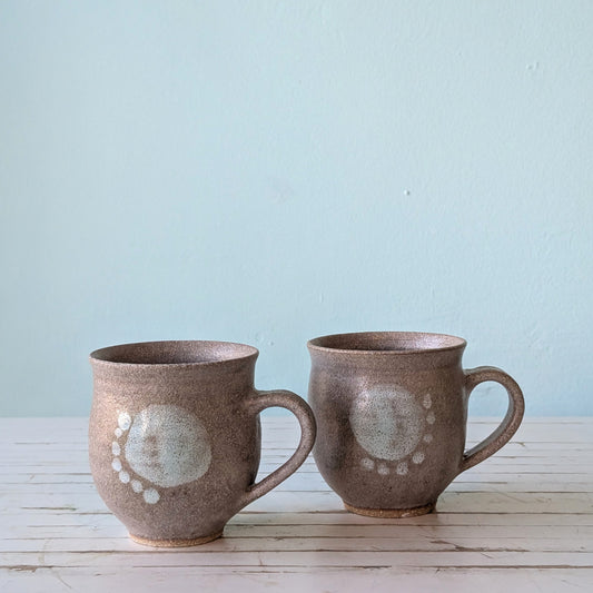 Front view of two brown stoneware mugs with blue/green abstract pattern on front. Mugs are sitting on a white table with a blue bakcground.