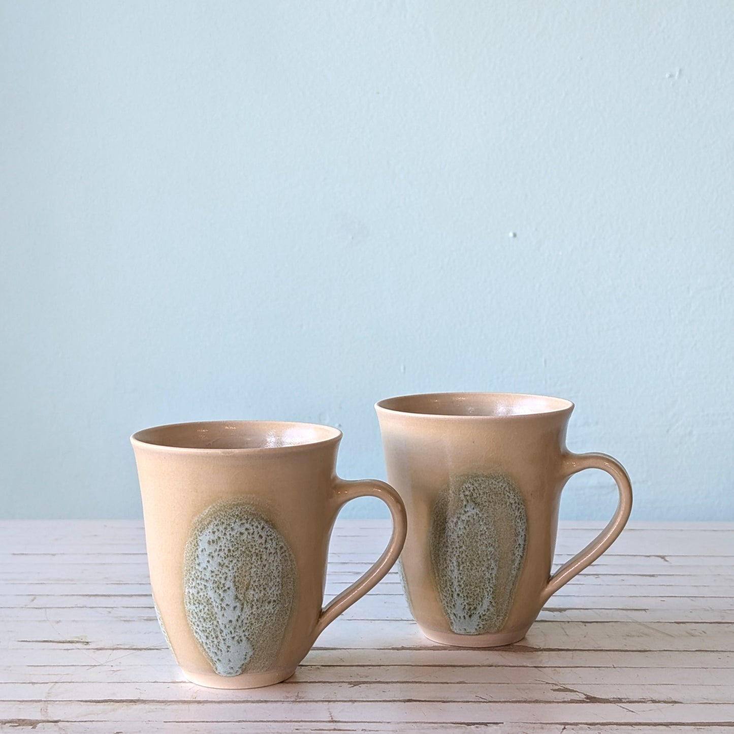 Front view of two mugs with handles on a white table with blue background. Mugs are beige stoneware with green brushstroke design.