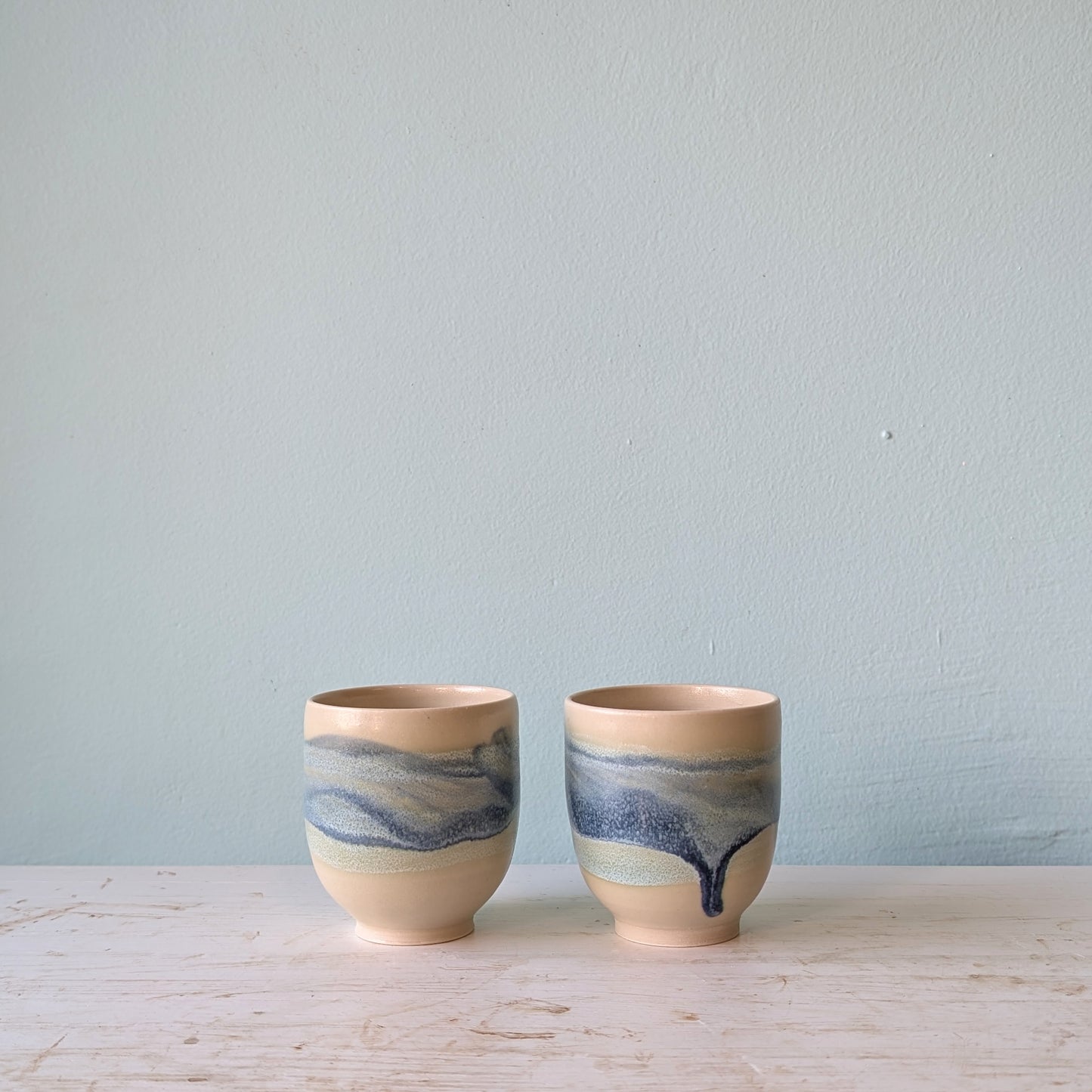 Two beige and blue ceramic cups on a white table against a blue wall.
