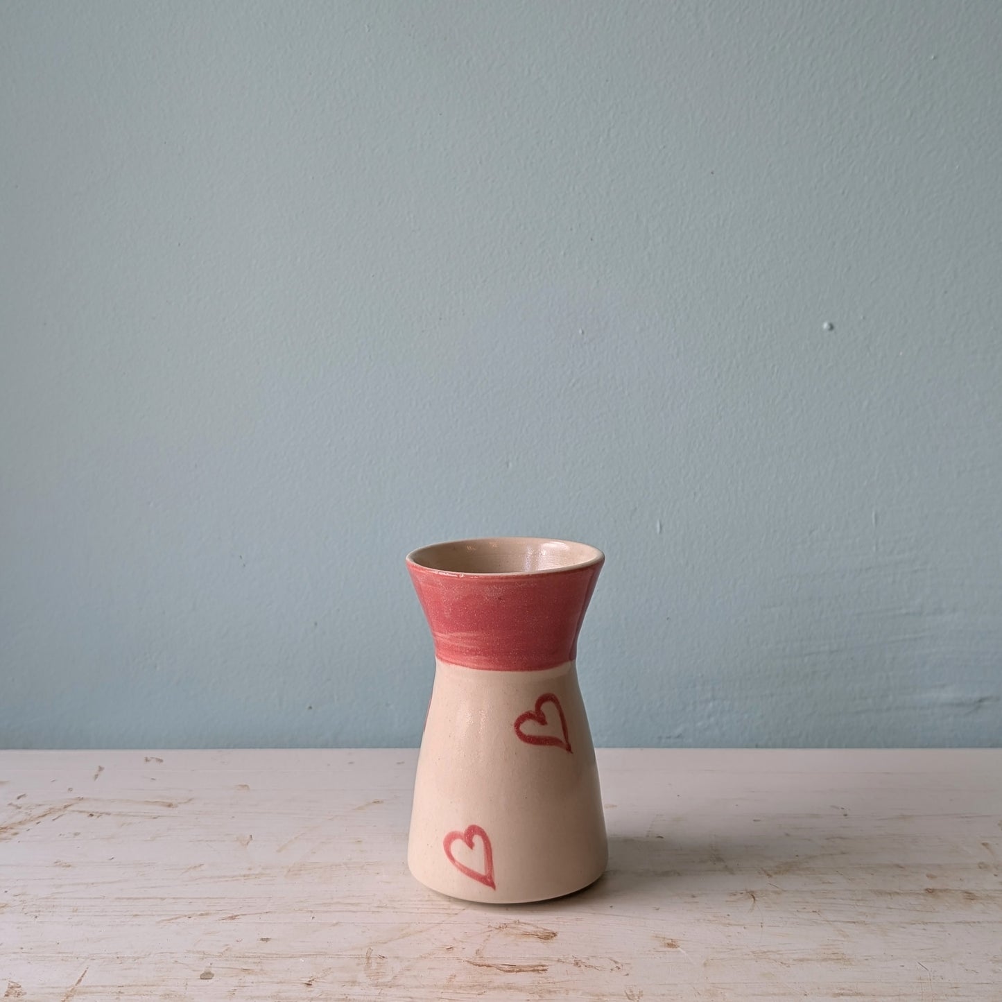 Pink and beige ceramic vase with see through pink hearts on a white table against a blue wall.