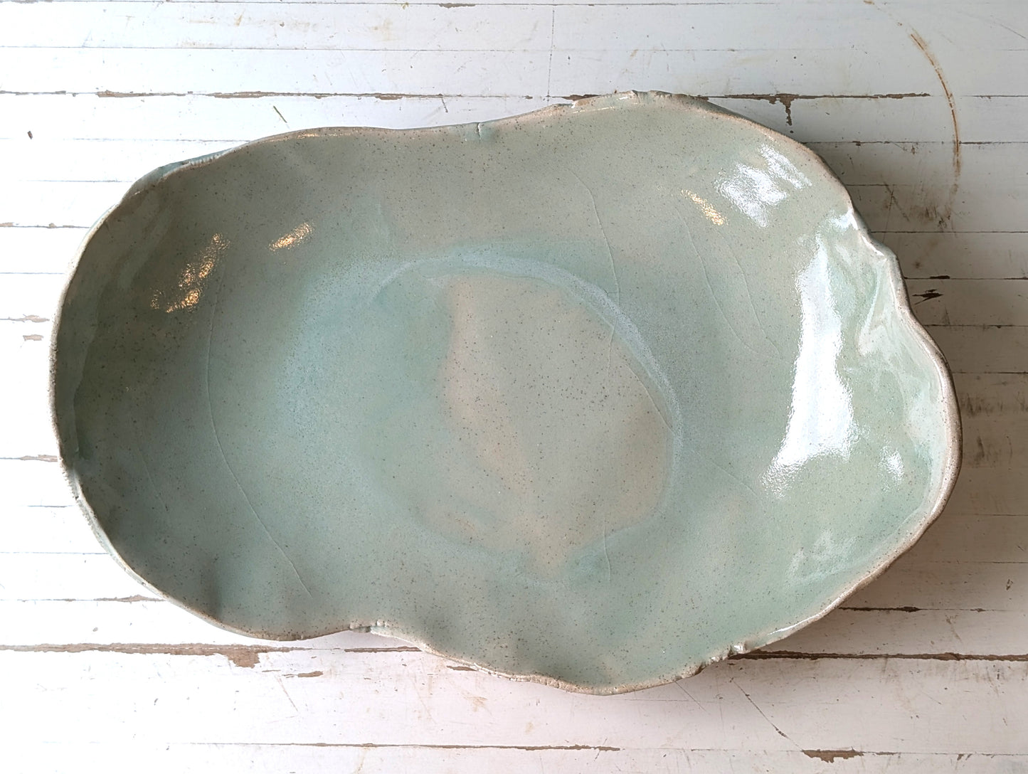 Top view of a freeform ceramic serving platter in light green, on a white wooden table.