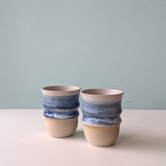 Two beige ceramic cups with dark blue glaze on a white table with a blue background.