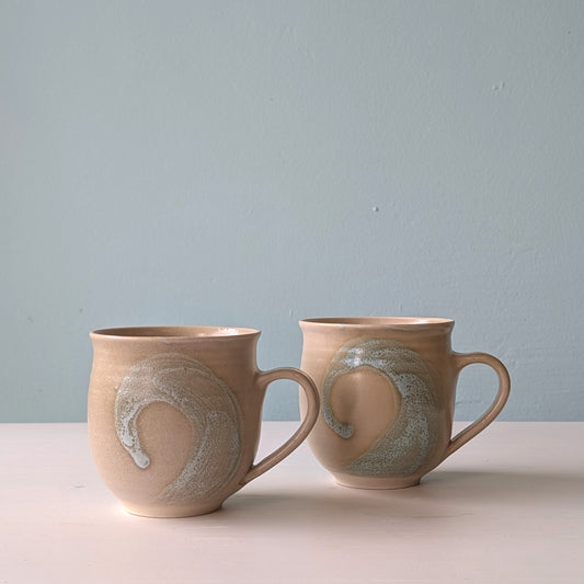 Two beige ceramic mugs with green wave design on a white table against a blue background.