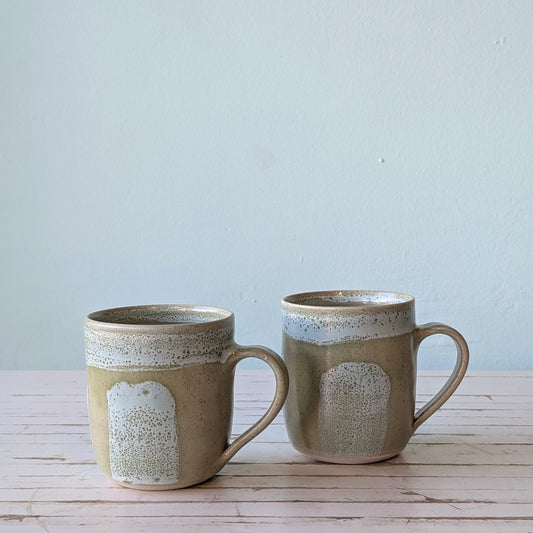 Front view of two green ceramic mugs with blue/green brushstroke pattern, sitting on a white table with a blue background.