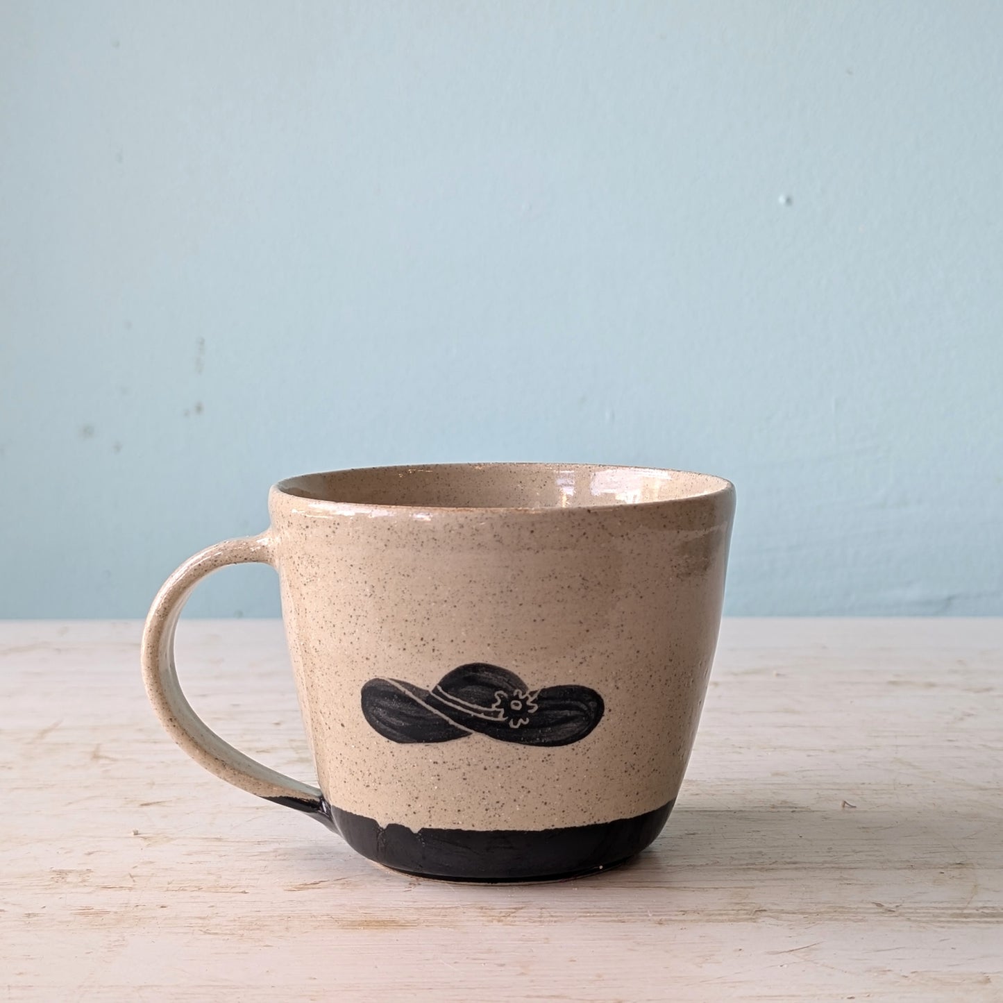 Beige ceramic mug with black hat design on a light wooden surface and light blue background