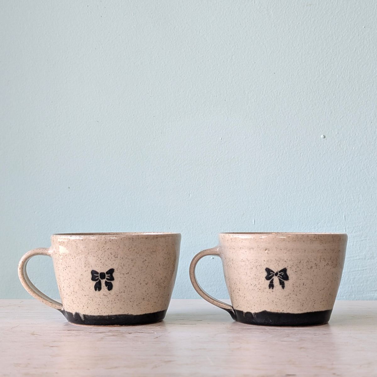 Two handmade ceramic mugs with speckled beige glaze, black bow motifs, and black footed bases, displayed side-by-side on a light wooden surface against a pale blue wall.