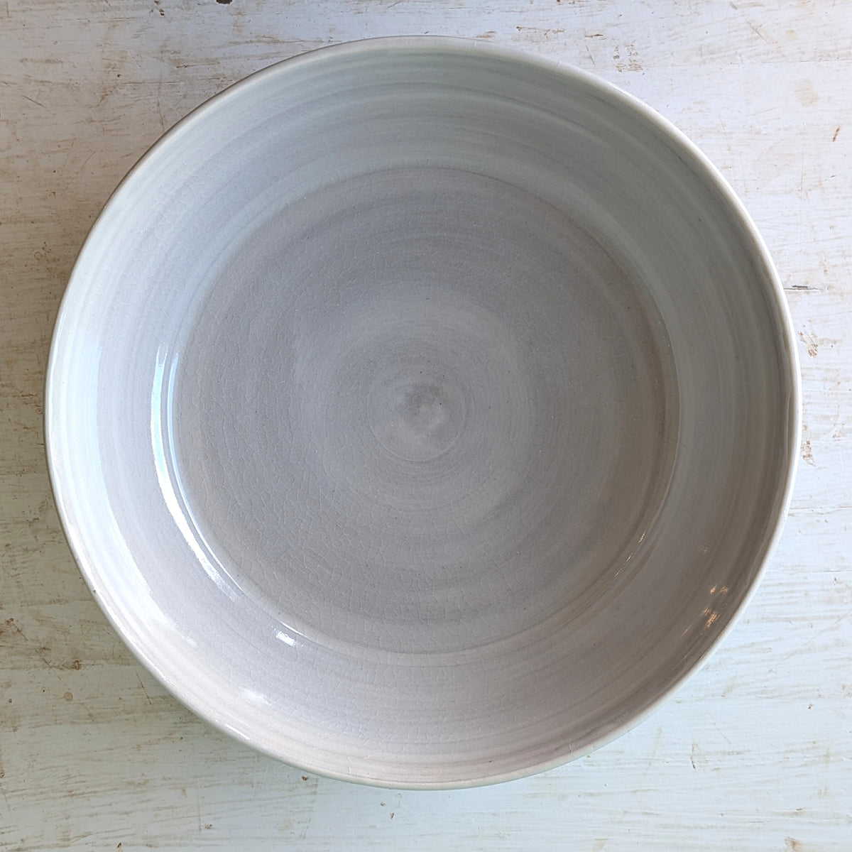 A round ceramic bowl with a wide rim, photographed from directly above on a distressed white wooden surface. The bowl has a smooth, glossy finish with a white glaze and a hand-painted white edge.