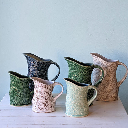 Front view of 6 Denise McDonald flannel flower pitchers in sizes large and small. Colours pictured are blue, green, teal and buff white.