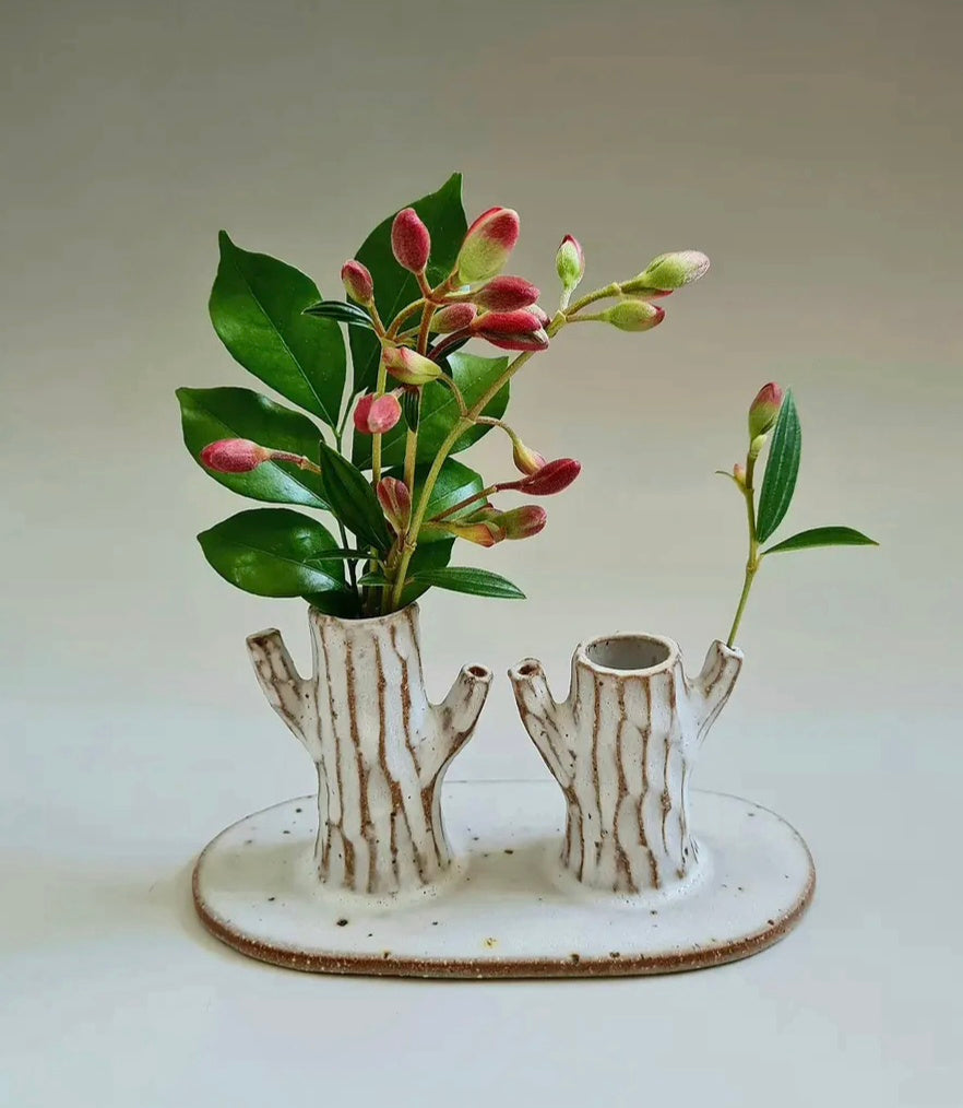 A ceramic two-bottle vase designed to look like tree stumps, with a textured off-white glaze and brown bark-like vertical striations. It holds green leaves and pinkish-red flower buds, set against a neutral light gray background.