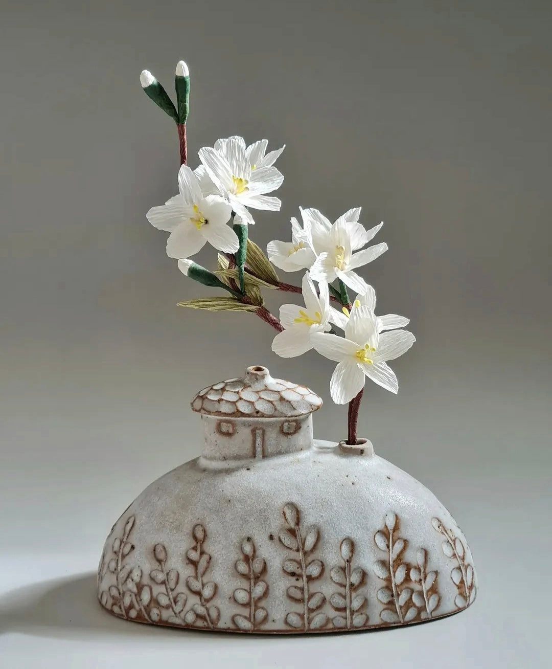 A ceramic bud vase shaped like a closed teacup with a small house on top, decorated with brown leaf patterns. It holds white paper flowers with green stems and buds, set against a neutral gray background.