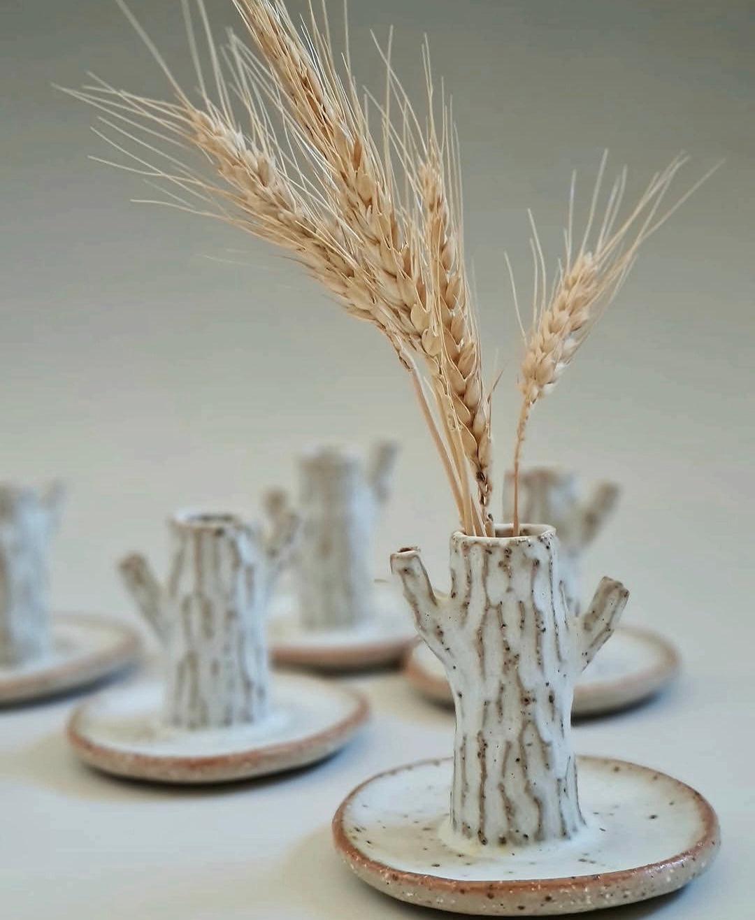 A ceramic tree-bud vase with a attached saucer, styled with dried wheat stalks, set against a softly blurred background of similar vases.
