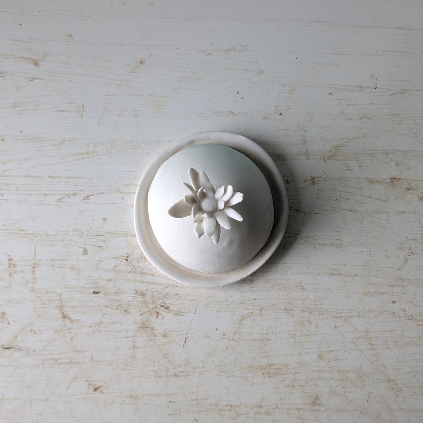 A small white ceramic lidded container or dish with a sculpted flower on top, photographed from above on a distressed white wooden surface.