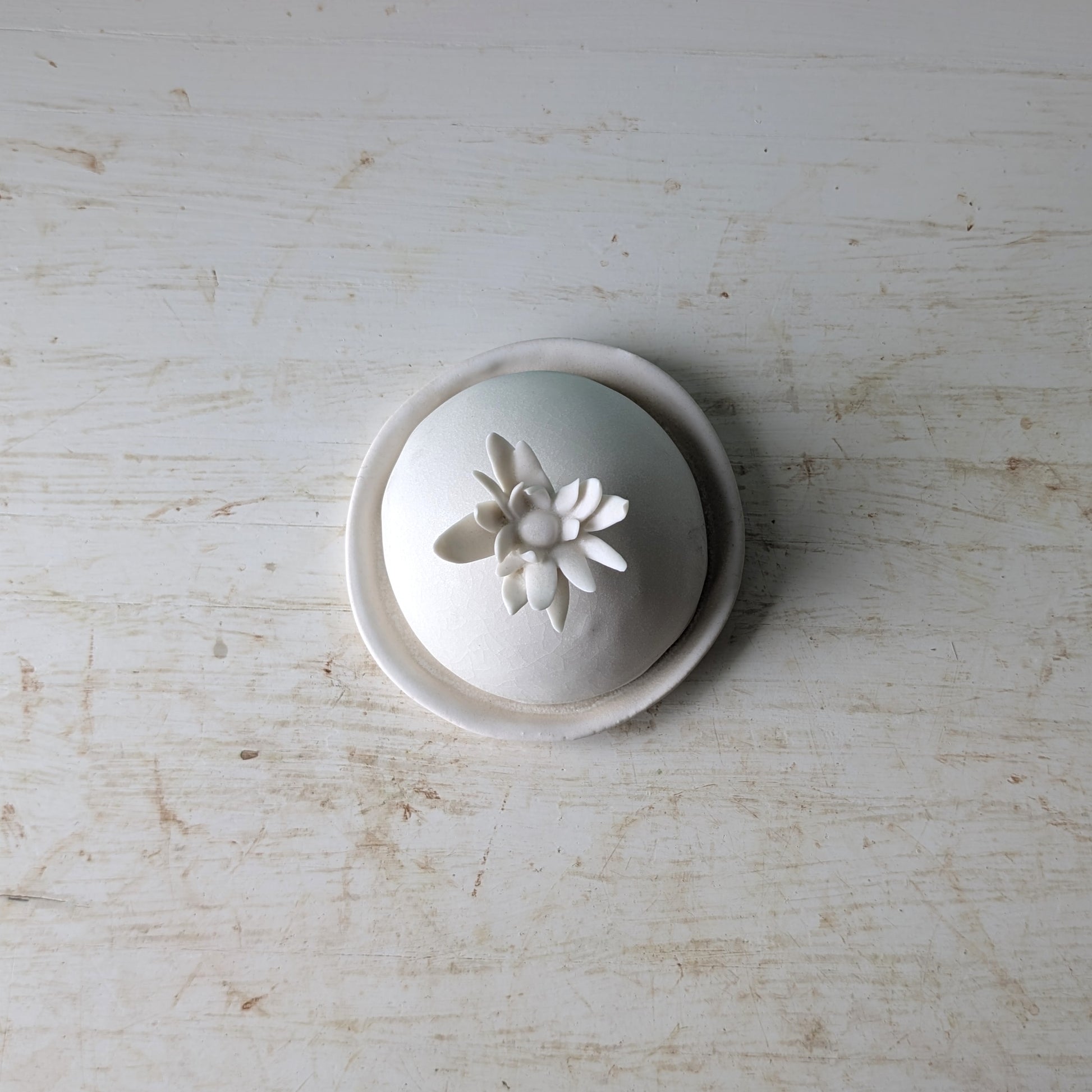 A small white ceramic lidded container or dish with a sculpted flower on top, photographed from above on a distressed white wooden surface.