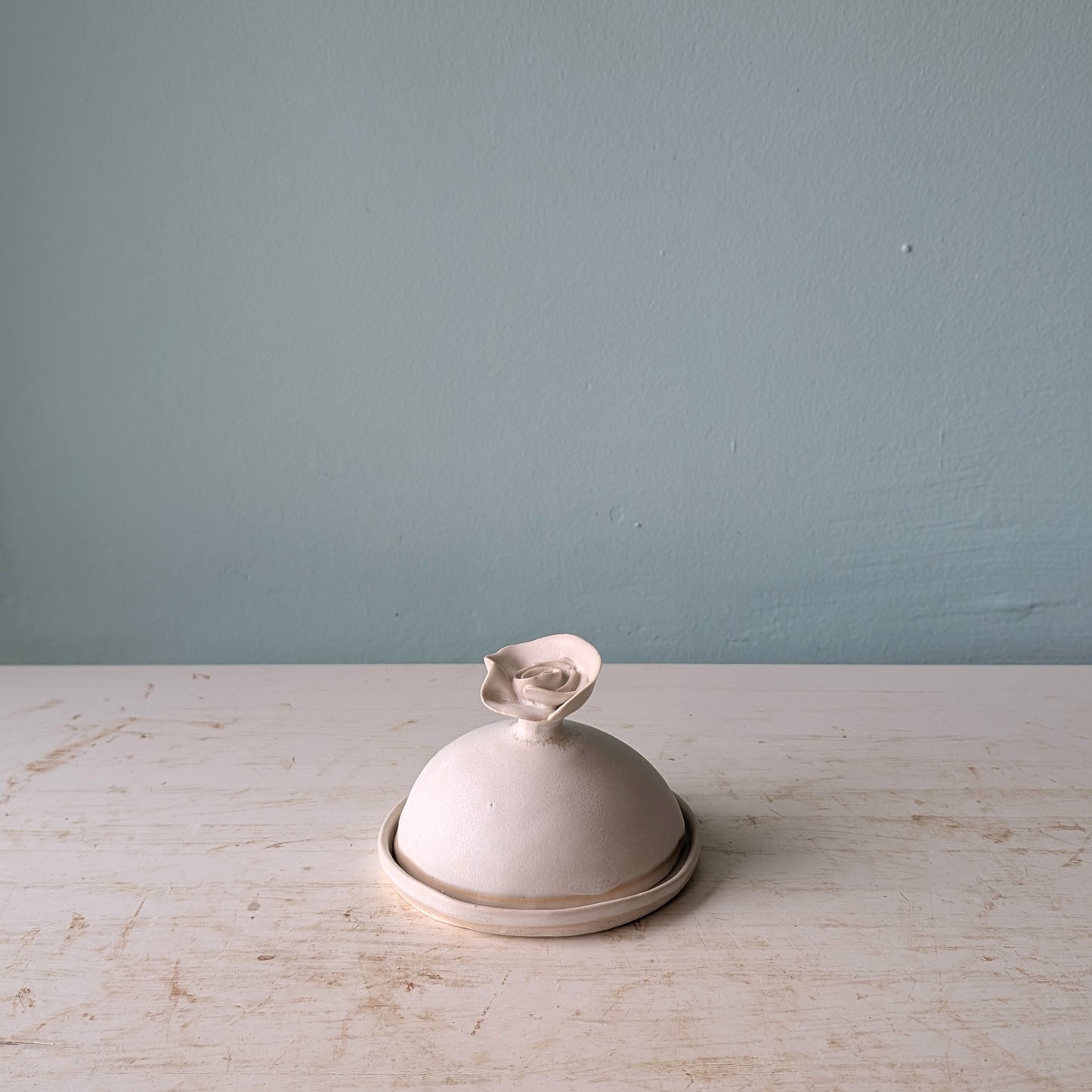 A small white ceramic butter dish with a flower-shaped handle, sitting on a distressed white wooden surface against a blue wall.