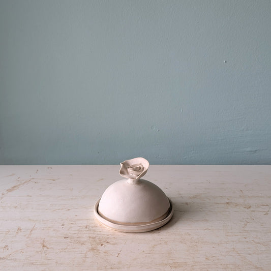 A small white ceramic butter dish with a flower-shaped handle, sitting on a distressed white wooden surface against a blue wall.