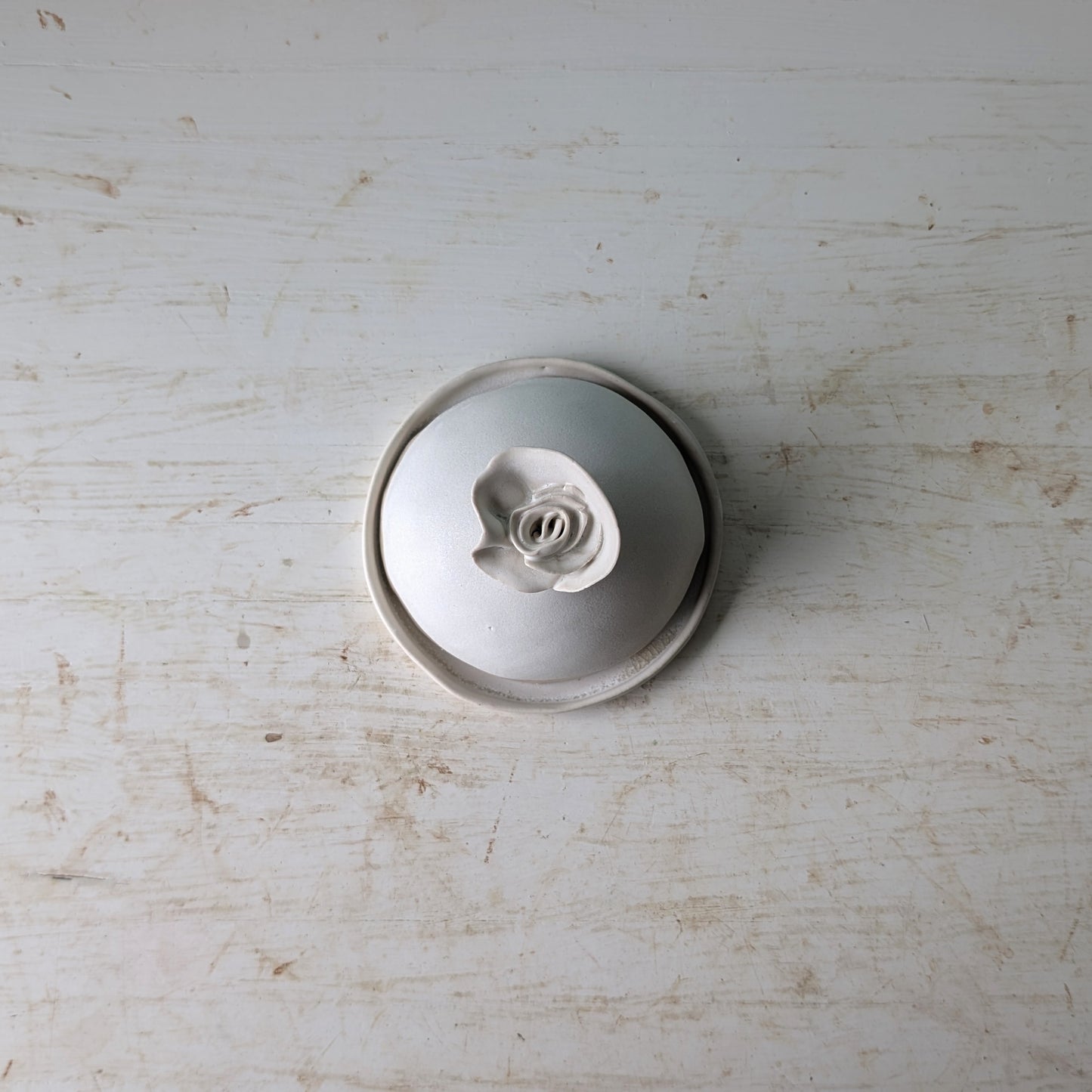 A small round white ceramic butter dish with a sculpted rose-shaped handle, photographed from above on a distressed white wooden surface.