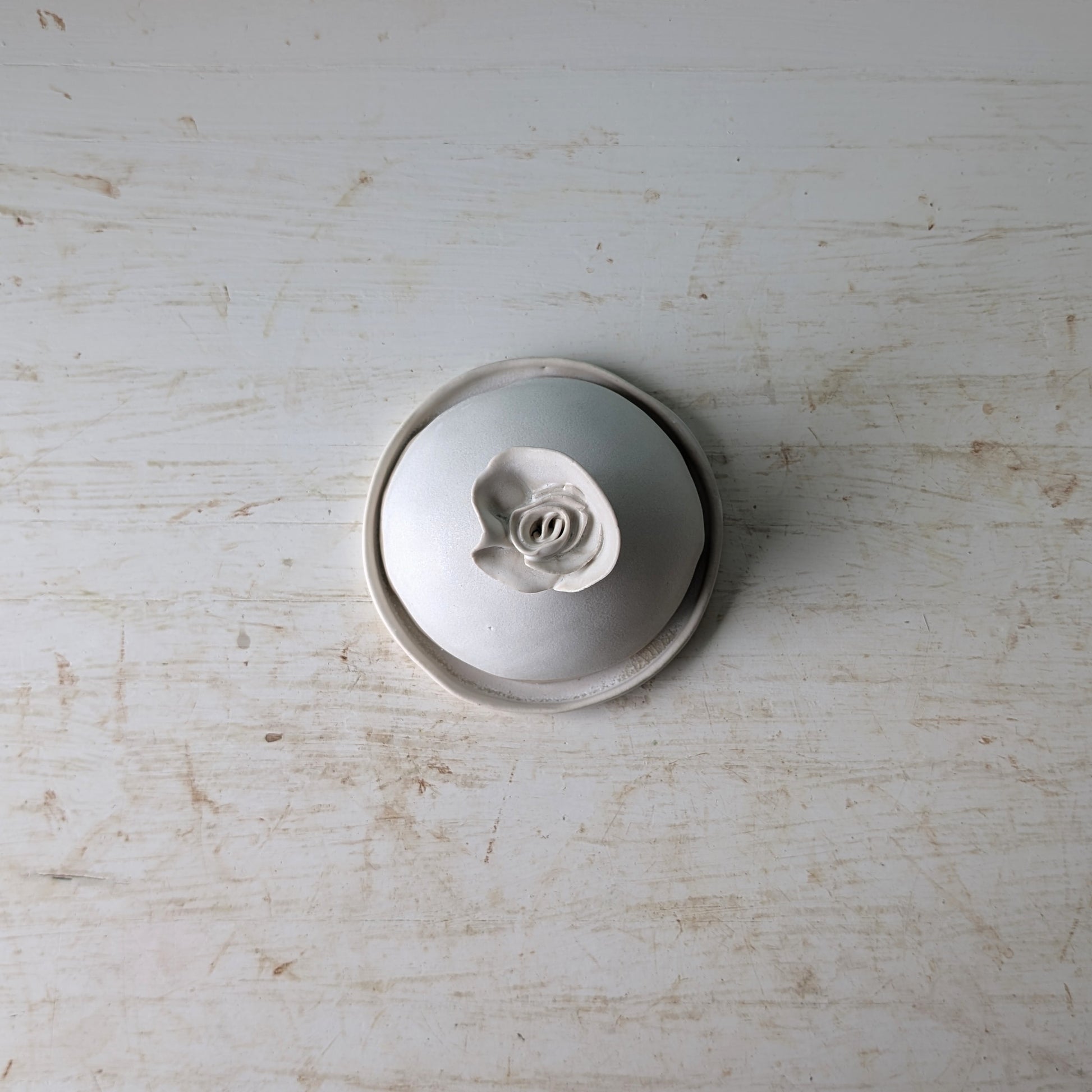 A small round white ceramic butter dish with a sculpted rose-shaped handle, photographed from above on a distressed white wooden surface.