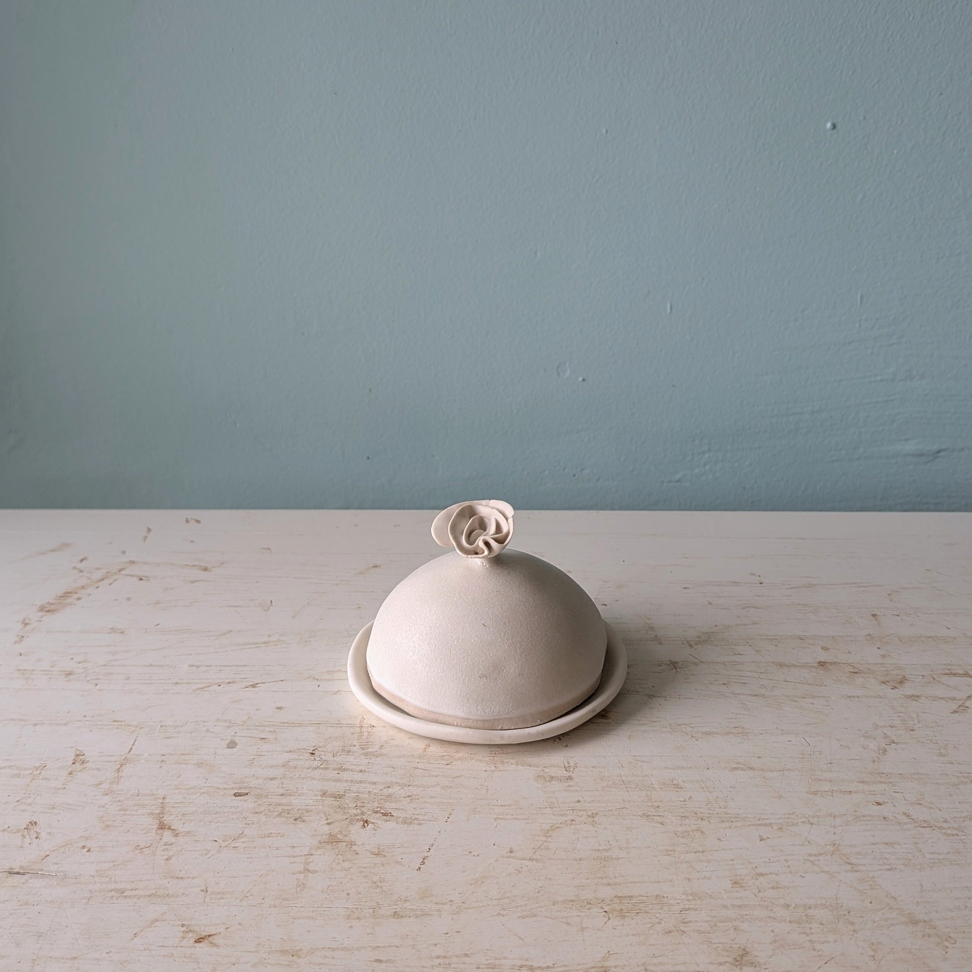 A small dome-shaped ceramic butter with a matching lid and a decorative flower-shaped knob, set on a white wooden surface against a blue wall.