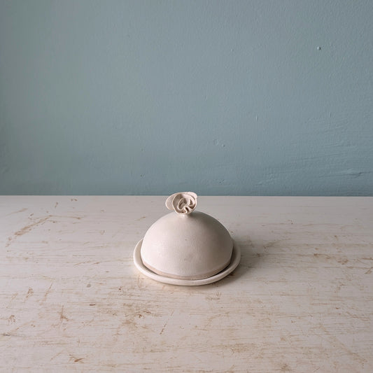 A small dome-shaped ceramic butter with a matching lid and a decorative flower-shaped knob, set on a white wooden surface against a blue wall.