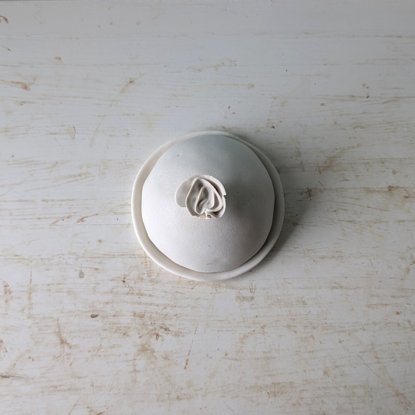 A small round ceramic butter dish with a lid, photographed from above on a distressed white wooden surface.