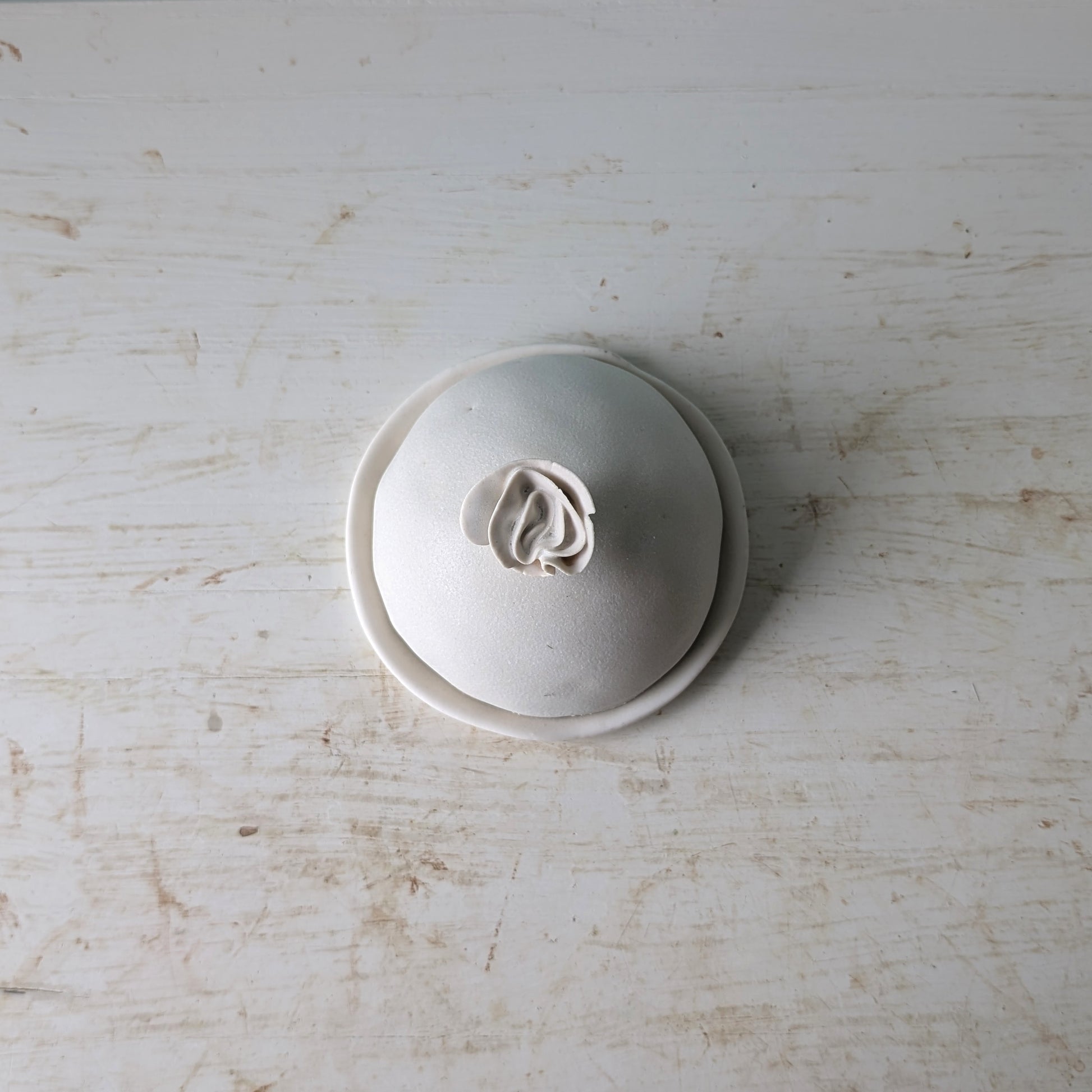 A small round ceramic butter dish with a lid, photographed from above on a distressed white wooden surface.