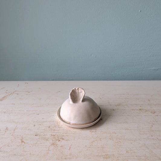 A small handmade ceramic lidded butter dish with a rounded lid and textured handle, sitting on a distressed white wooden surface against a muted blue-gray wall.
