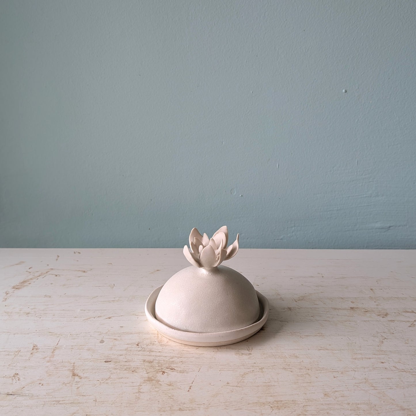 White ceramic butter dish with domed lid and flower shaped handle, on a white wooden table against a blue wall.