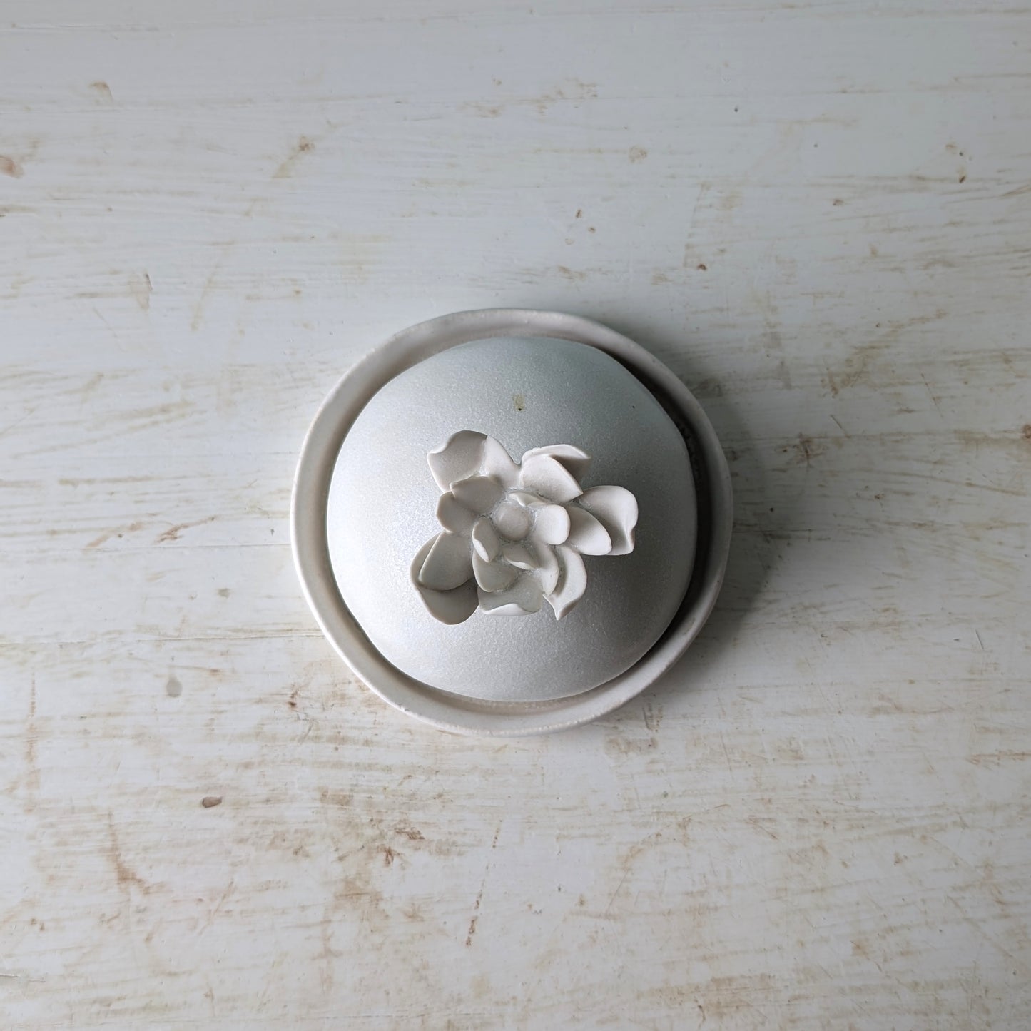 A white ceramic lidded butter dish with a sculptural flower-shaped knob, photographed from above on a distressed white wooden surface.
