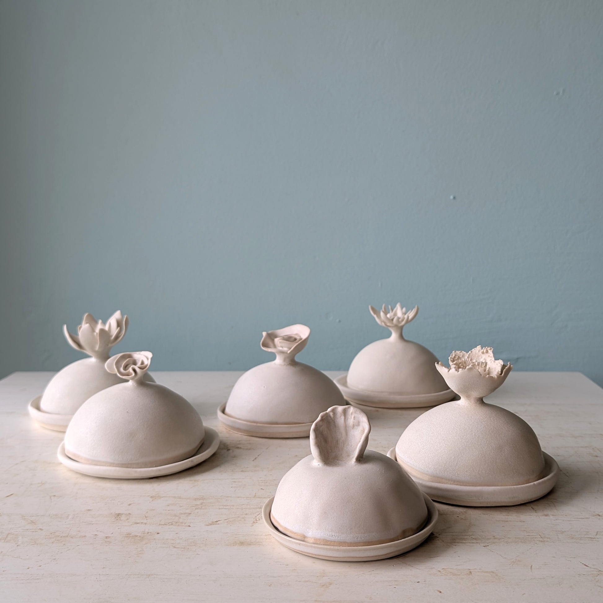 A small matte ceramic lidded butter dish with a sculptural flower-like lid and matching saucer, set on a light wood surface against a muted blue wall.