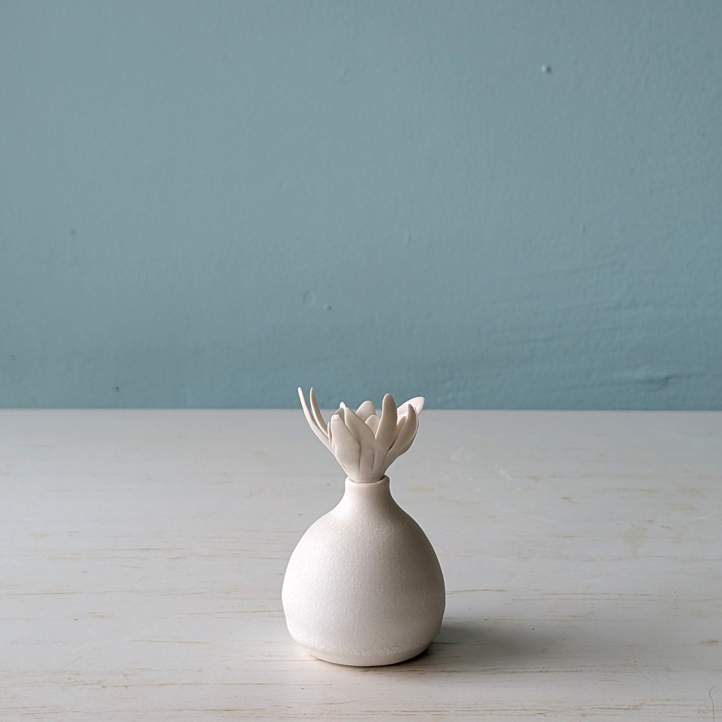 White ceramic bottle with a unique design on a light wooden surface against a blue wall.