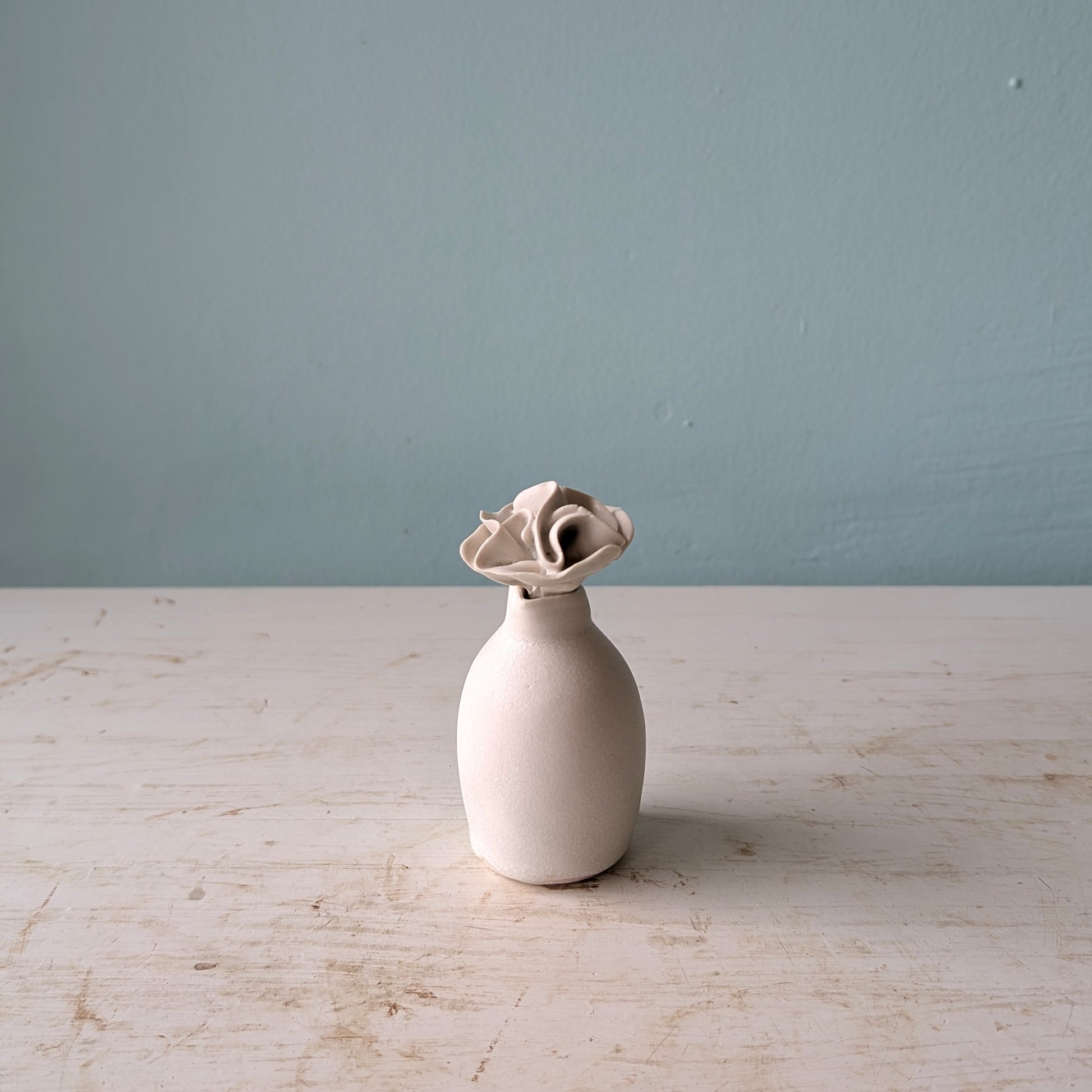 A small matte ceramic vase with a flared, flower-like top, standing on a light distressed tabletop against a blue wall.