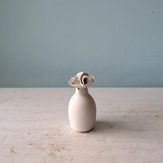 A small matte ceramic vase with a flared, flower-like top, standing on a light distressed tabletop against a blue wall.