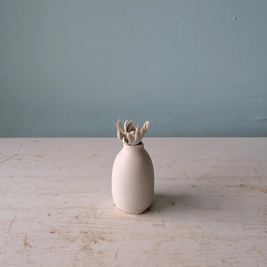 A small matte ceramic vase with a flared, flower-like top, standing on a light distressed tabletop against a blue wall.