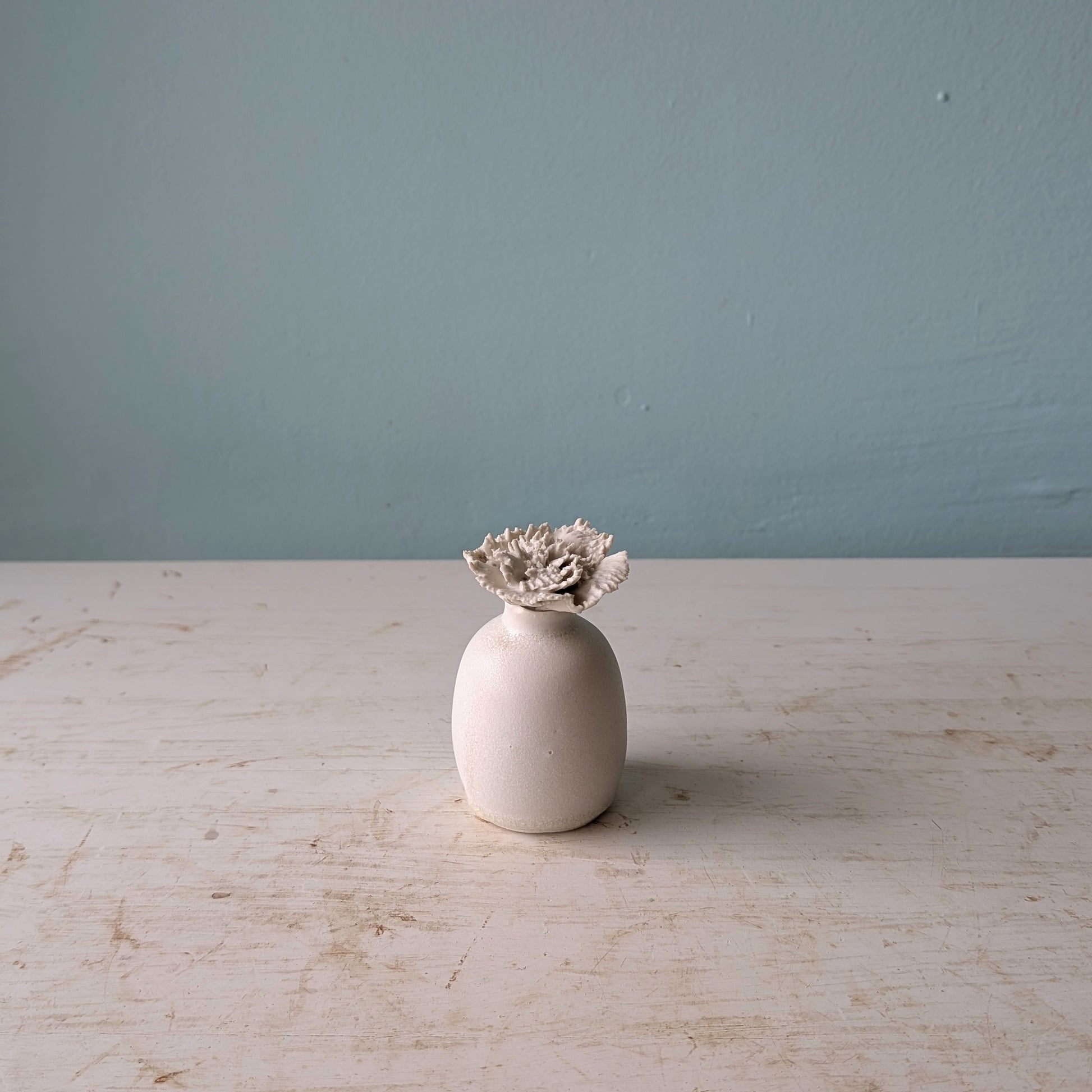 A small matte ceramic vase with a textured, sculptural flower bud emerging from the top, set on a distressed white wooden table against a blue wall.