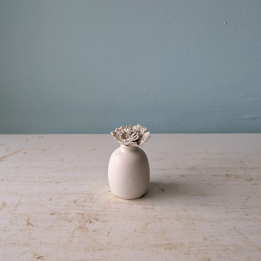 A small matte ceramic vase with a textured, sculptural flower bud emerging from the top, set on a distressed white wooden table against a blue wall.